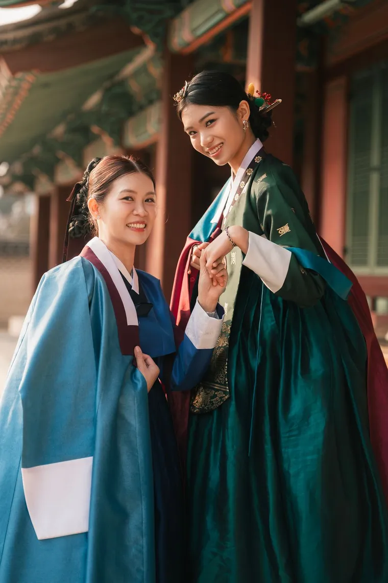 Two friends in blue and teal hanbok making finger hearts under a palace roof