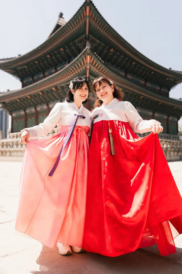 Two friends in pink and red hanbok holding out their skirts on the palace steps