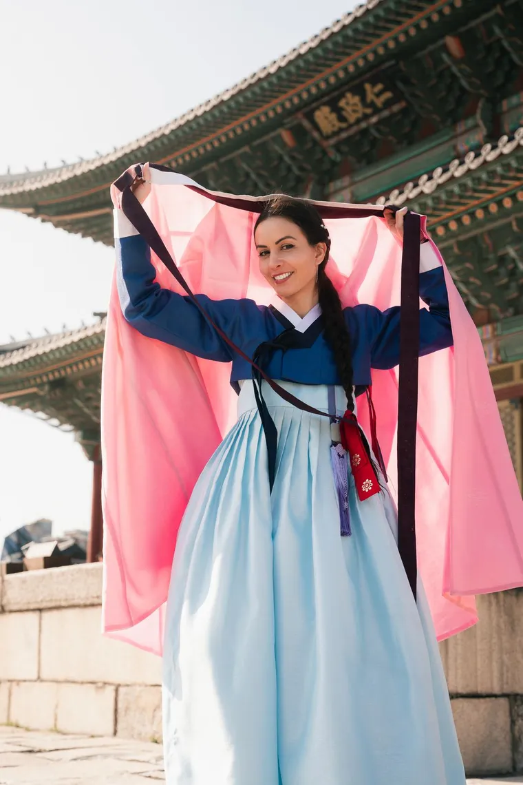 Woman in a light blue hanbok smiling with a pink shawl raised above her head