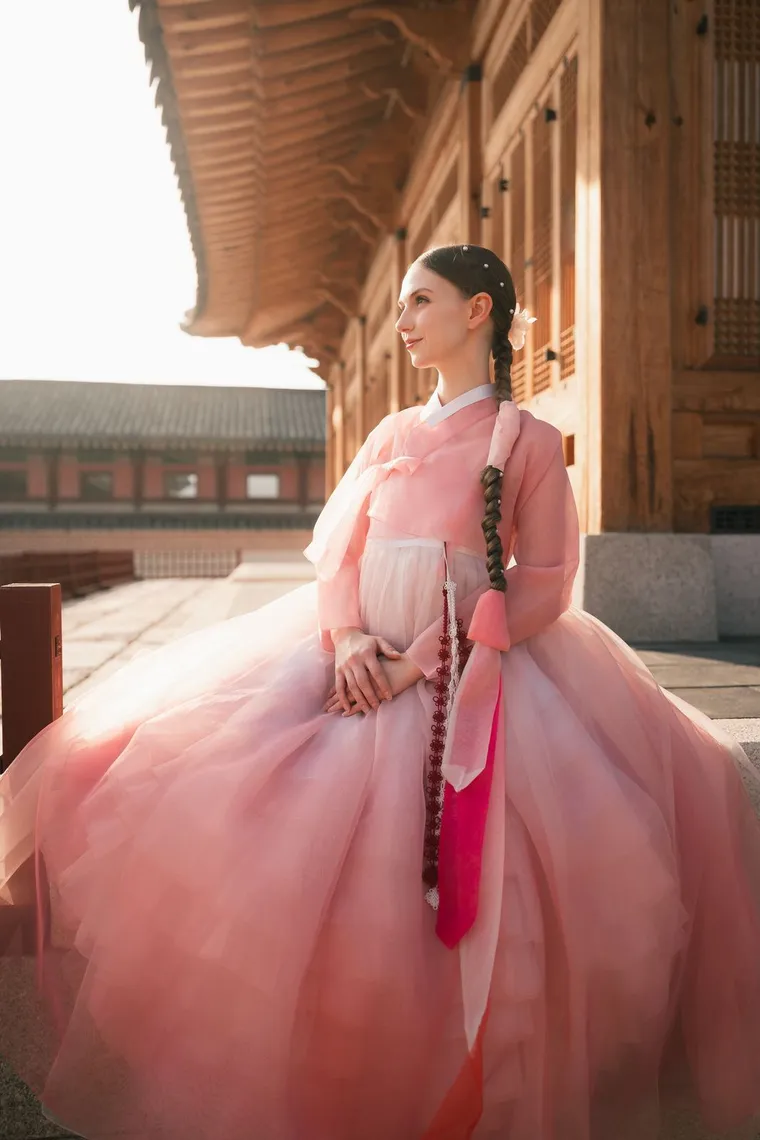 Woman in a pink hanbok with a long braid standing in a sunlit corridor