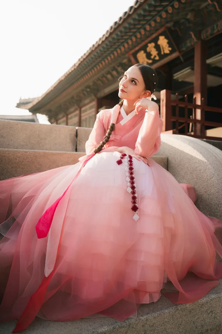 Woman in a pink hanbok seated on a palace stair, skirt spread wide