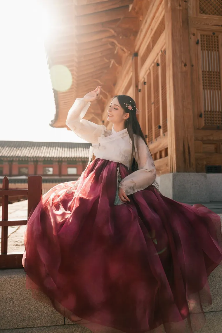 Woman in a crimson hanbok skirt seated on a palace stair, hand raised