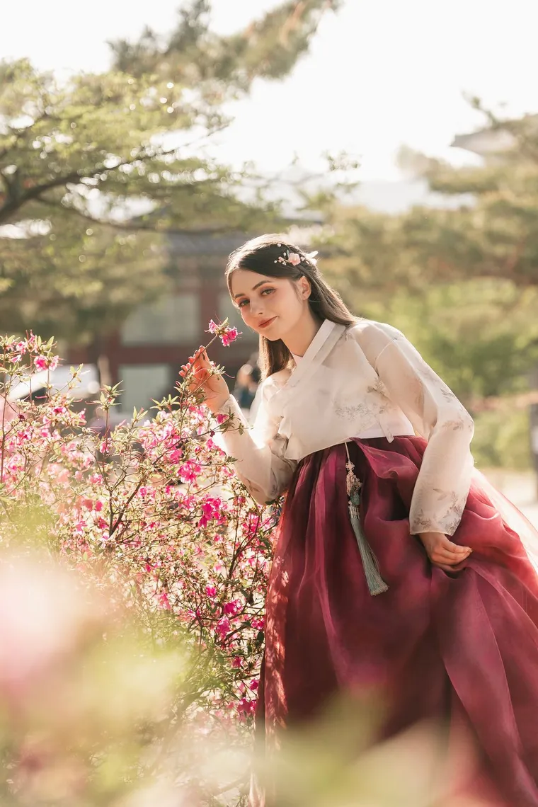 Woman in a crimson hanbok reaching toward azalea blossoms
