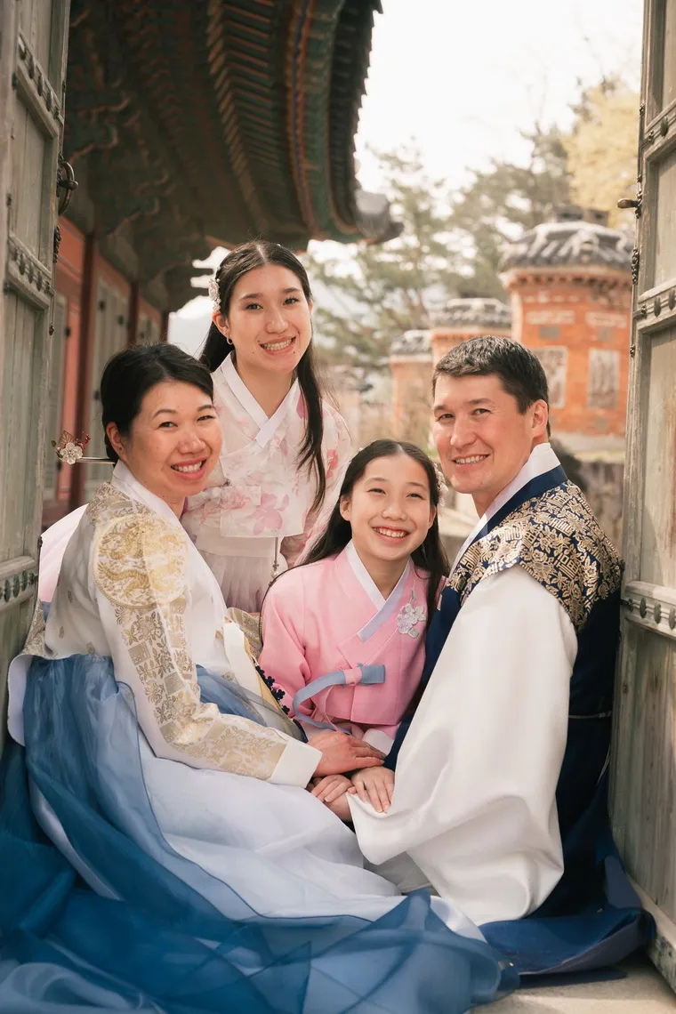 Family of four in hanbok smiling together beside a palace hall