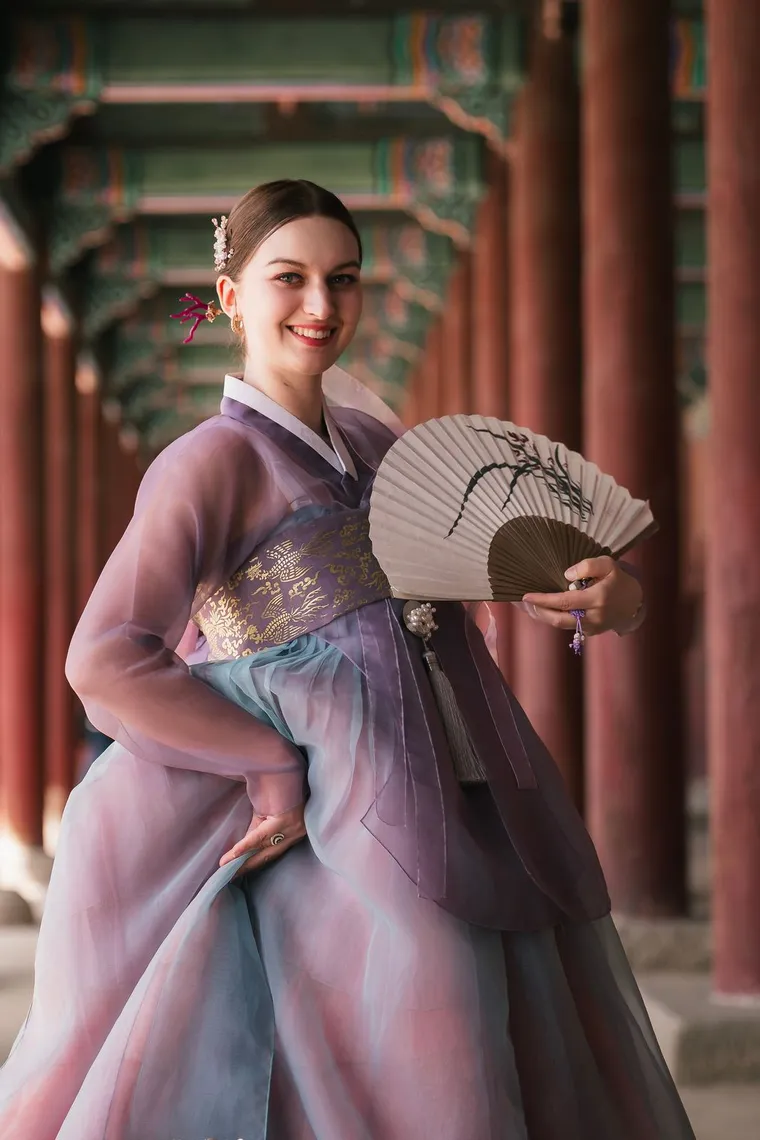 Woman in a lilac hanbok smiling with a folding fan in a red-columned corridor