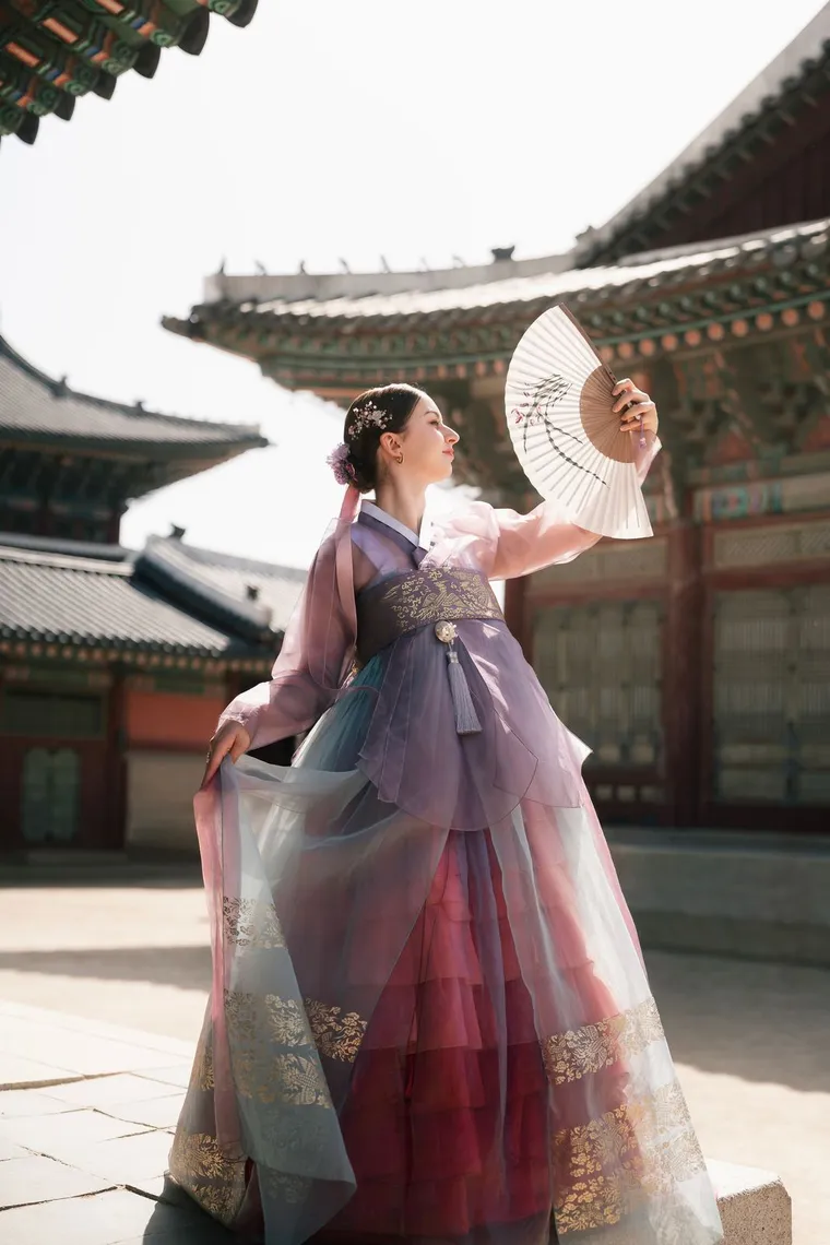 Woman in a lilac hanbok raising a folding fan in a palace courtyard